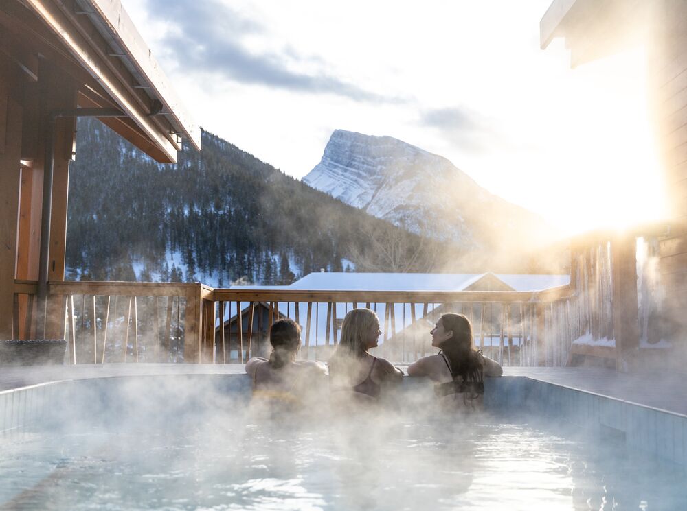 Three women hang out in the houtdoor hot tub at the Moose Hotel in Banff National Park with Mt. Rundle in the background.